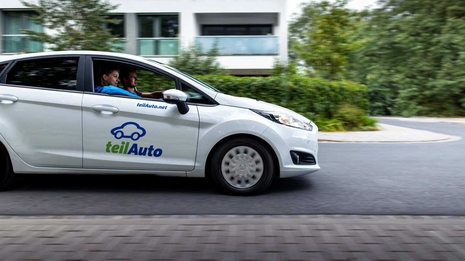 Ein Auto mit teilAuto-Logo fährt durch ein Wohngebiet.