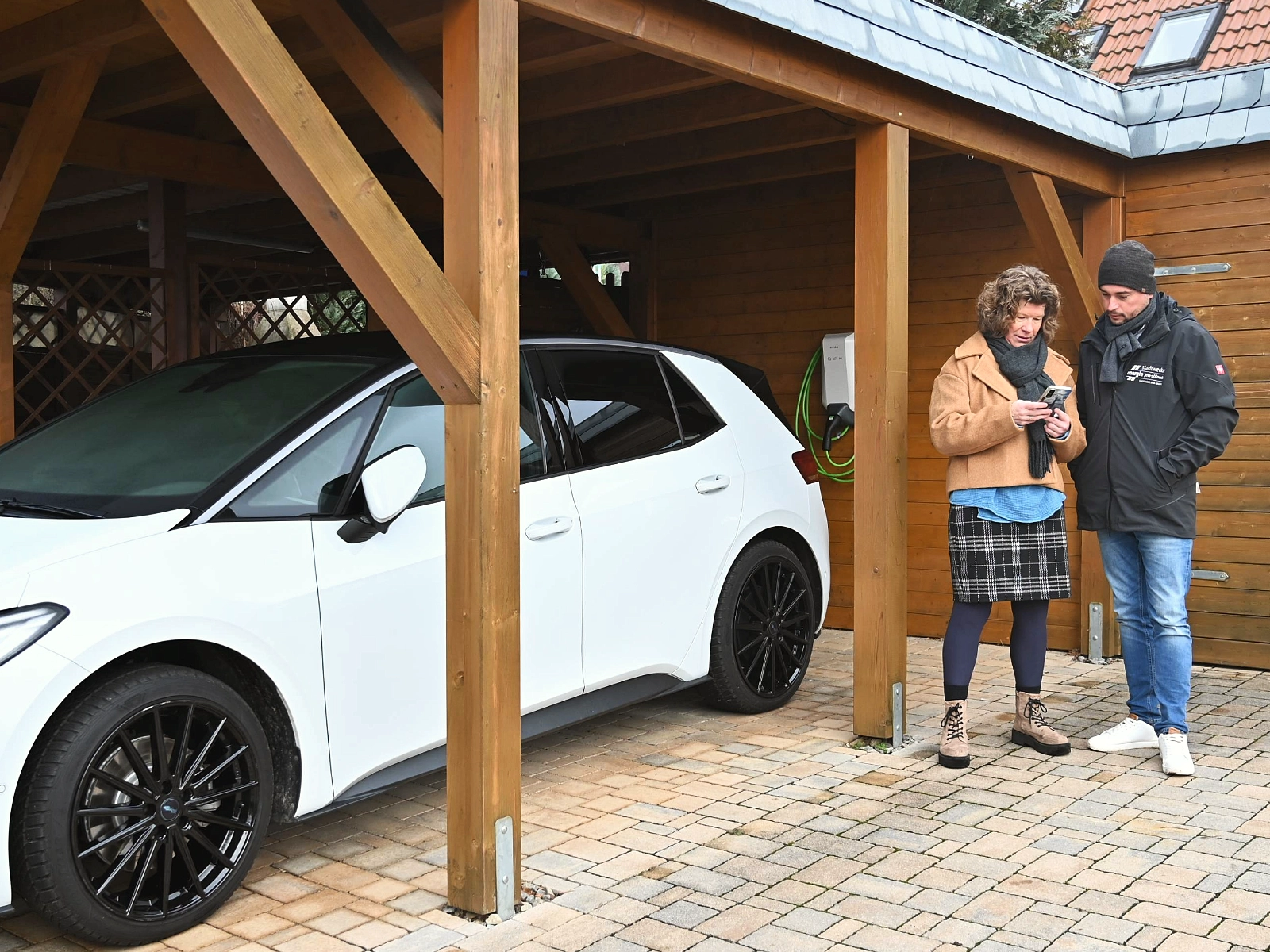 Katja Biertümpfel und Dennis Beyermann über ein Handy beugend im Gespräch, dahinter Wallbox und E-Auto.