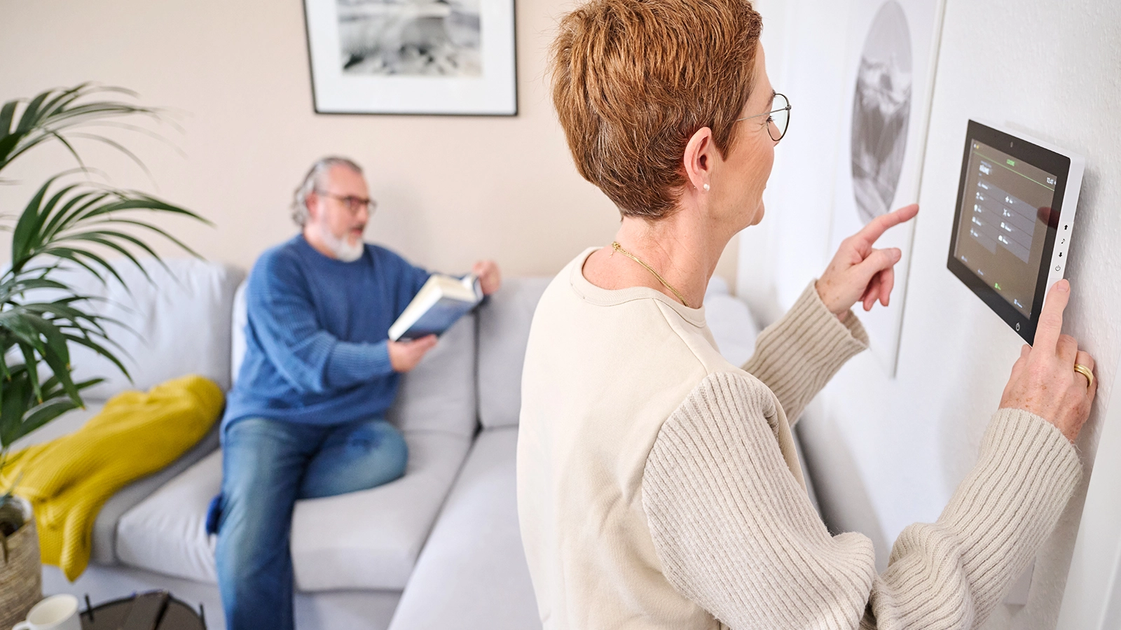 Ein Mann sitzt lesend auf dem Sofa, während eine Frau im Smarten Quartier ein Tablet bedient.