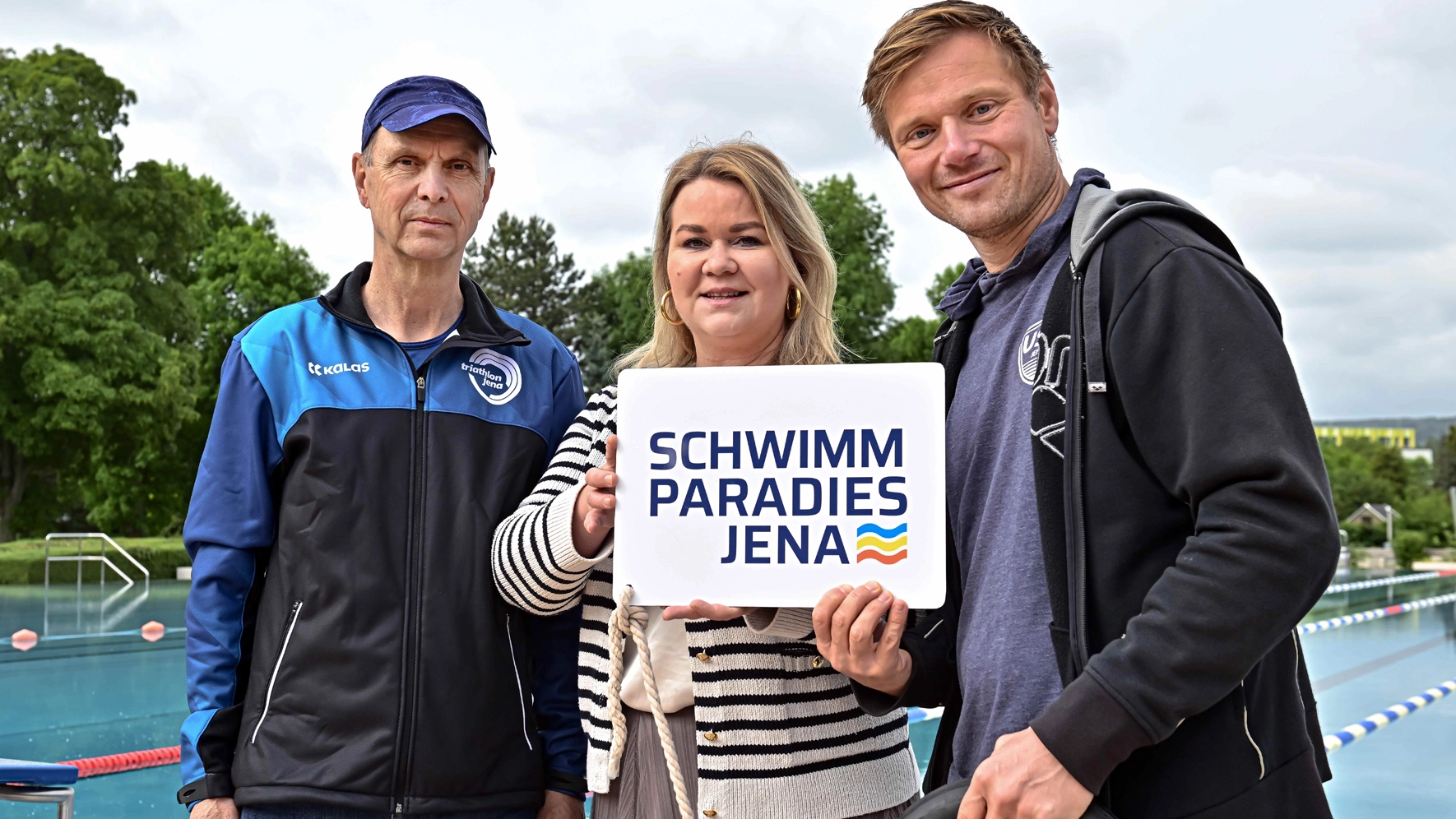 Drei Personen stehen vor einem Wasserbecken mit einem Schild in der Hand 