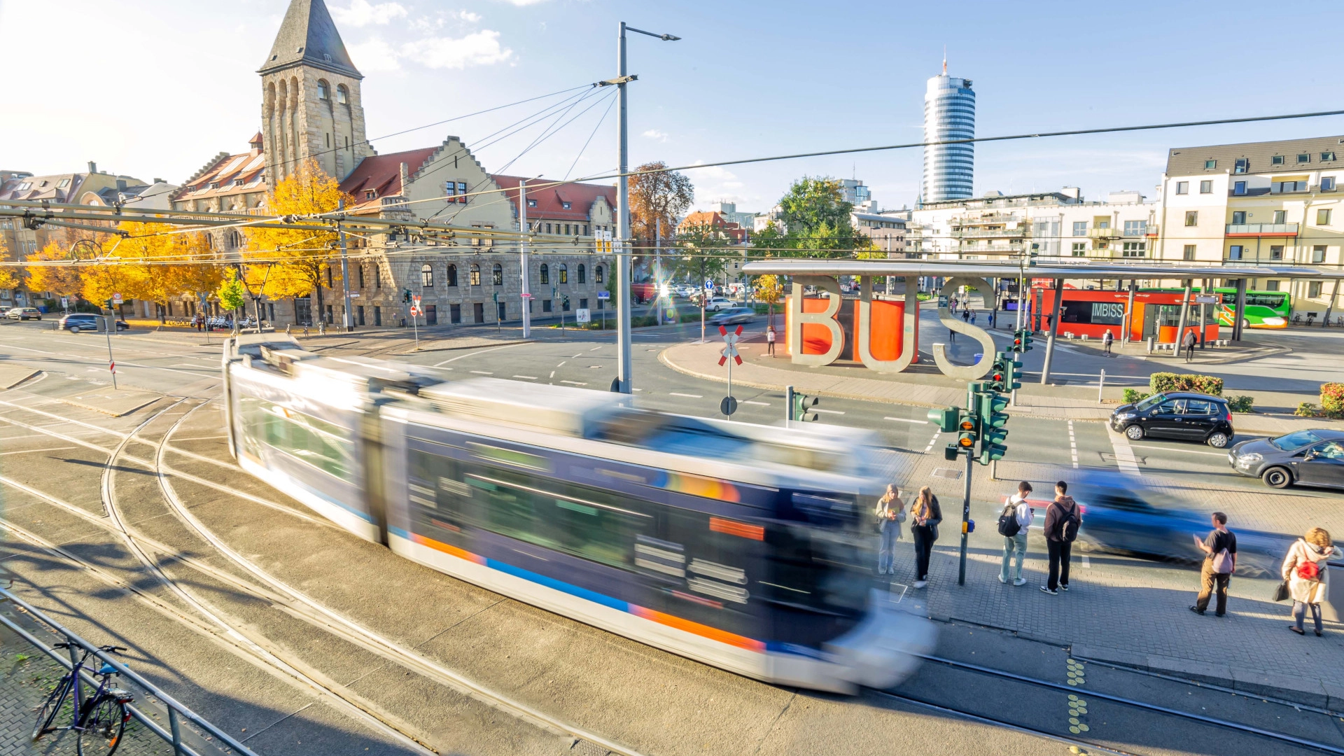 Der Busbahnhof in Jena mit einer fahrenden Straßenbahn im Vordergrund. Im Hintergrund das Volksbad.