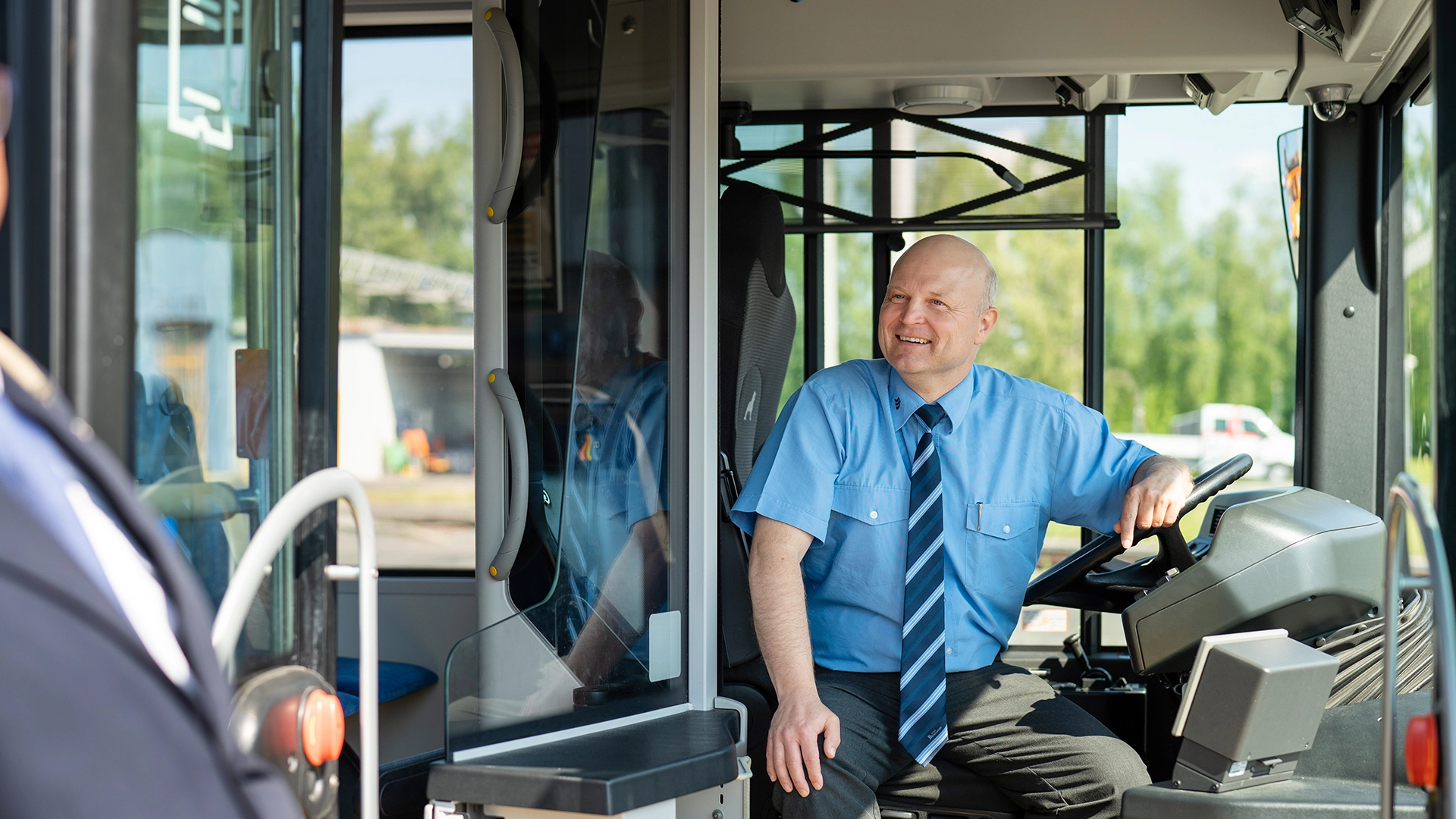 Ein Busfahrer sitzt lächelnd auf seinem Fahrersitz und schaut aus dem Bus. 