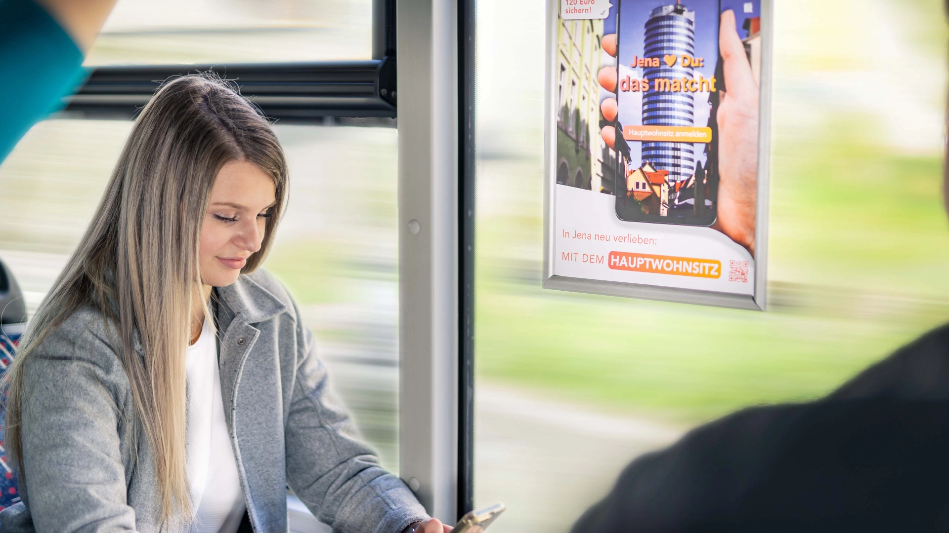 Eine Frau sitzt mit Handy im Bus und neben ihr ist Plakatwerbung zu sehen.
