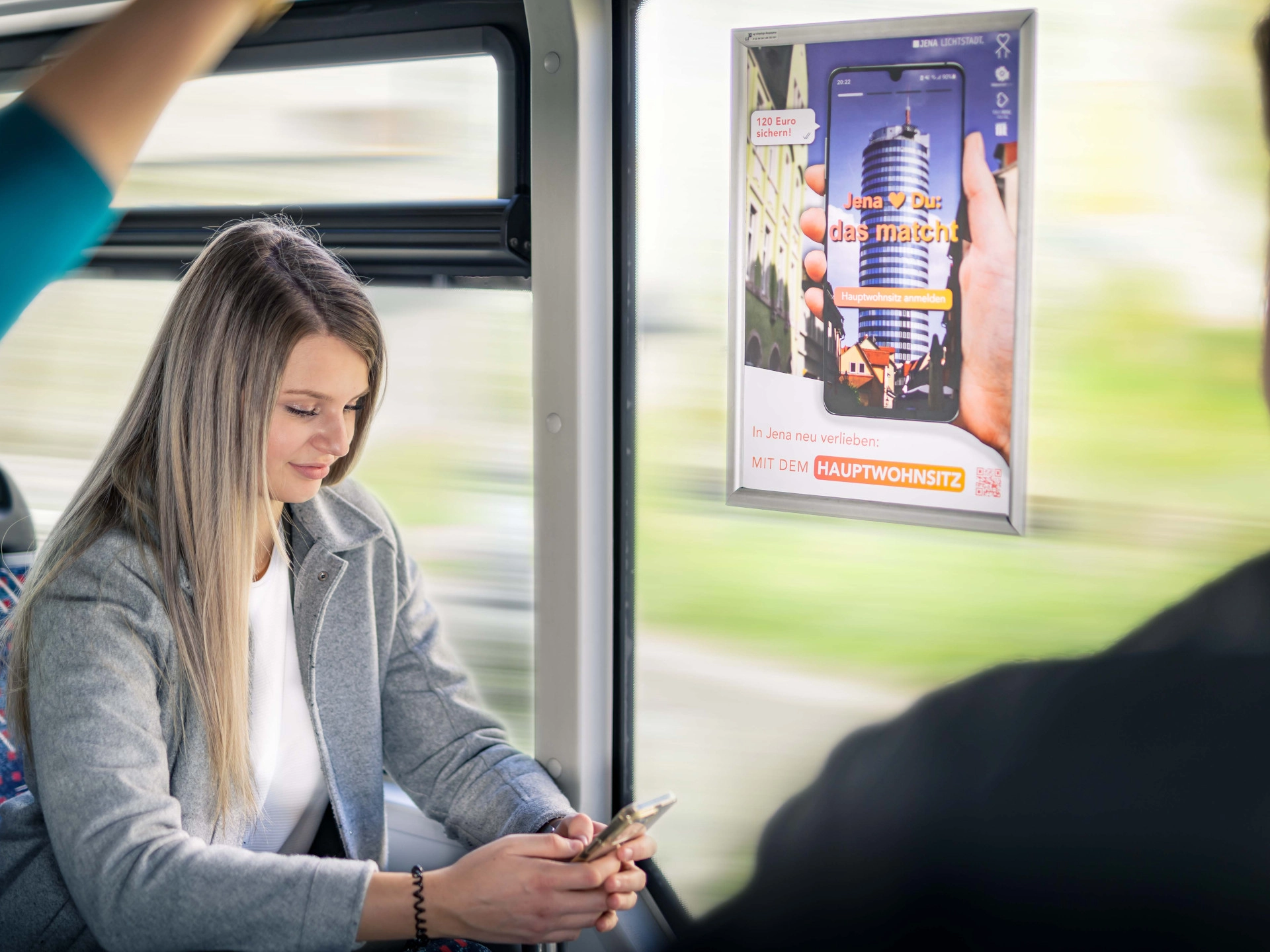 Eine Frau sitzt mit Handy im Bus und neben ihr ist Plakatwerbung zu sehen. Eine Frau sitzt mit Handy im Bus und neben ihr ist Plakatwerbung zu sehen.