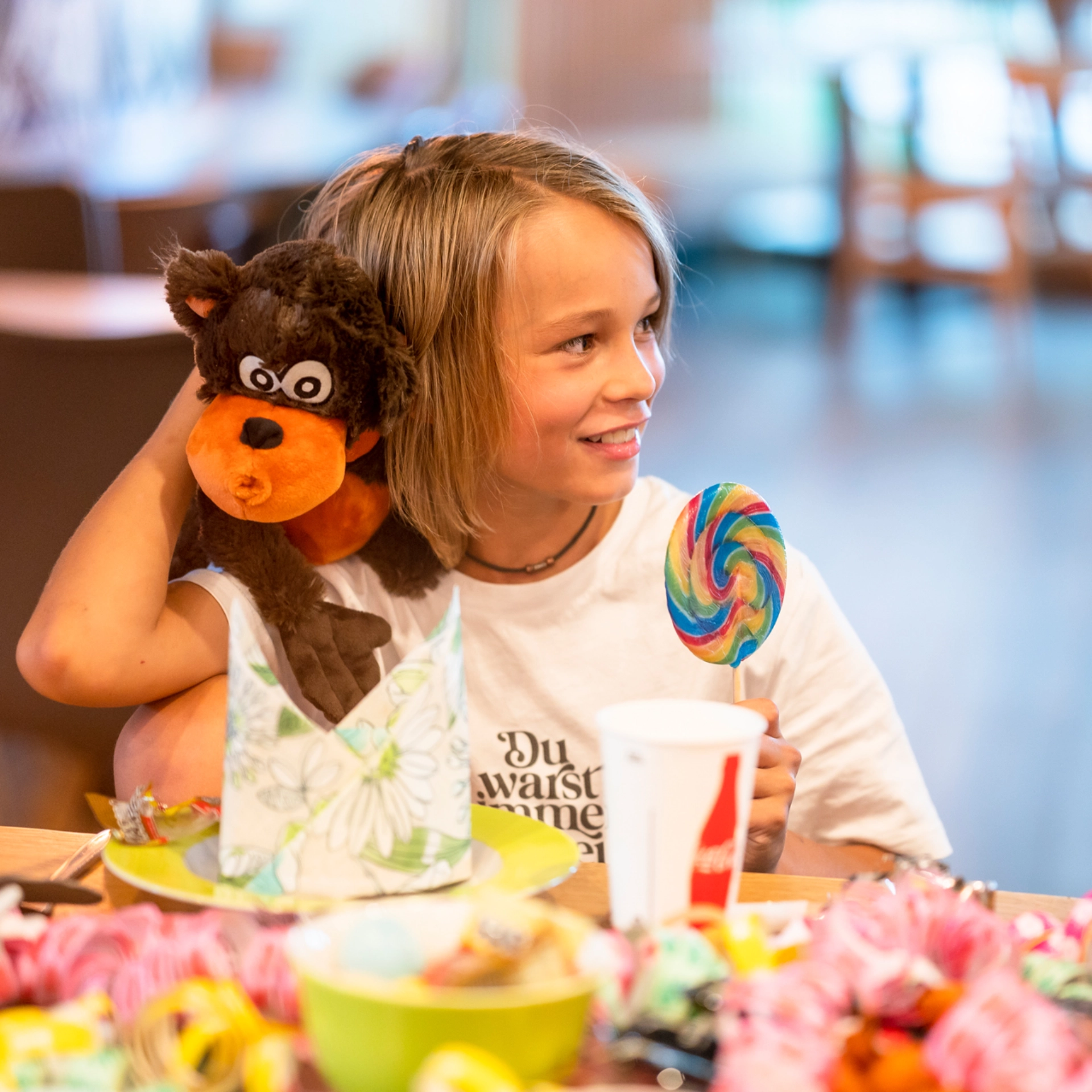 Ein Kind sitzt mit einem Kuscheltier und einem Lolly an einem Tisch