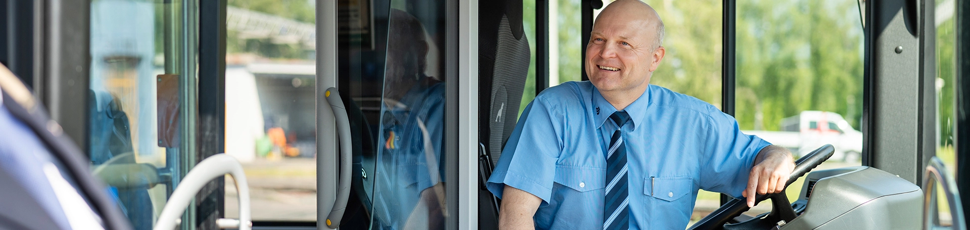 Ein Busfahrer sitzt lächelnd auf seinem Fahrersitz und schaut aus dem Bus. 