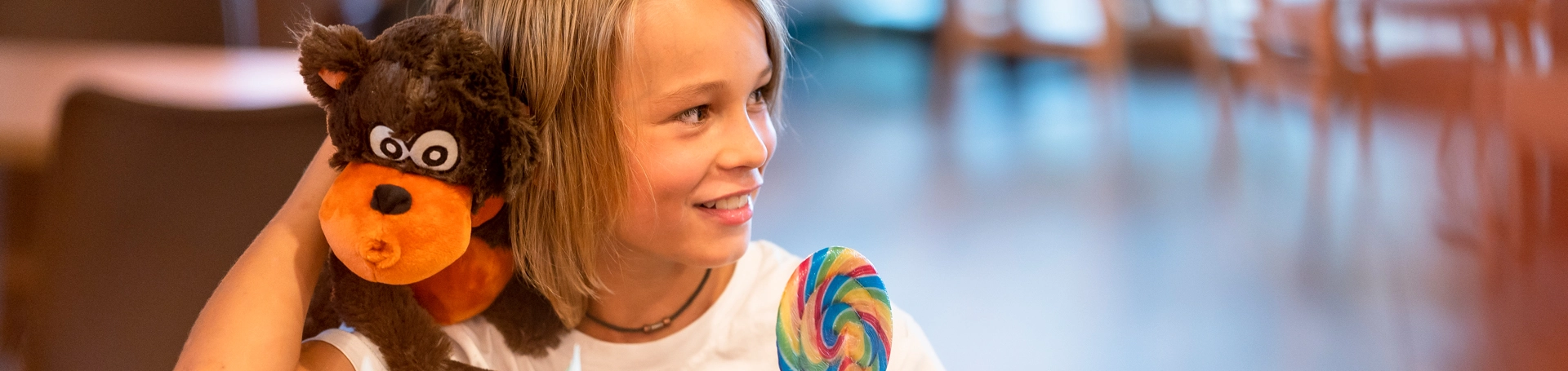 Ein Kind sitzt mit einem Kuscheltier und einem Lolly an einem Tisch