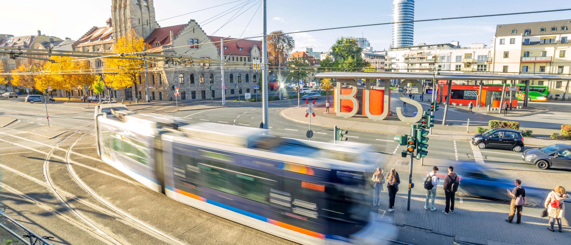 Der Busbahnhof in Jena mit einer fahrenden Straßenbahn im Vordergrund. Im Hintergrund das Volksbad.
