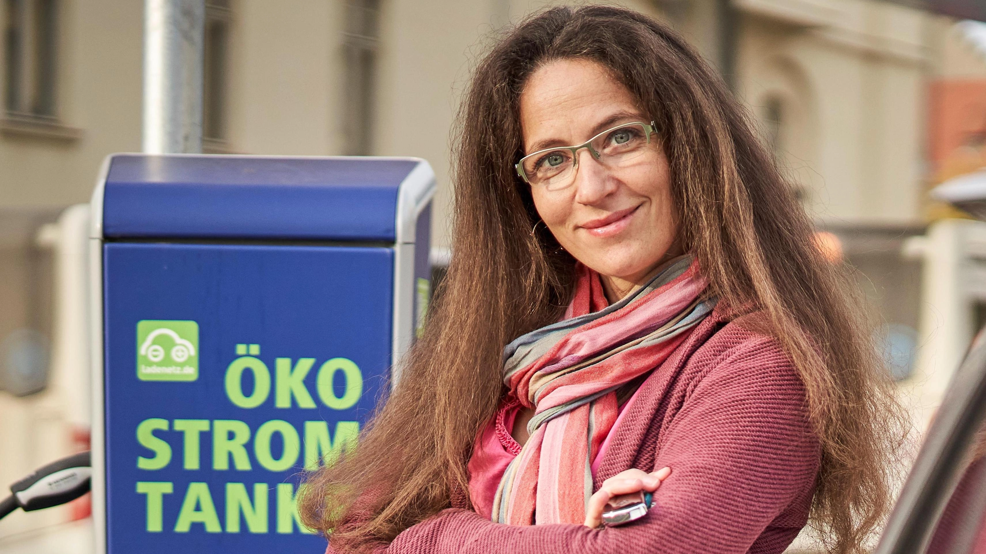 Eine E-Autofahrerin steht zufrieden vor einer Öko Strom Tanke