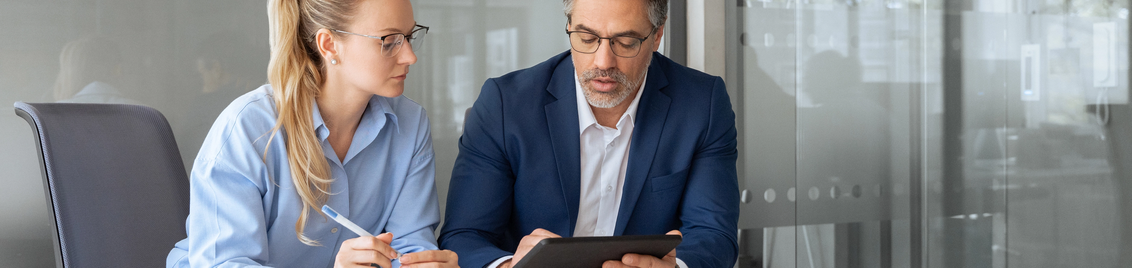 Eine Junge Frau und ein Mann unterhalten sich in einem Büro sitzend und schauen gemeinsam auf ein tablet. 