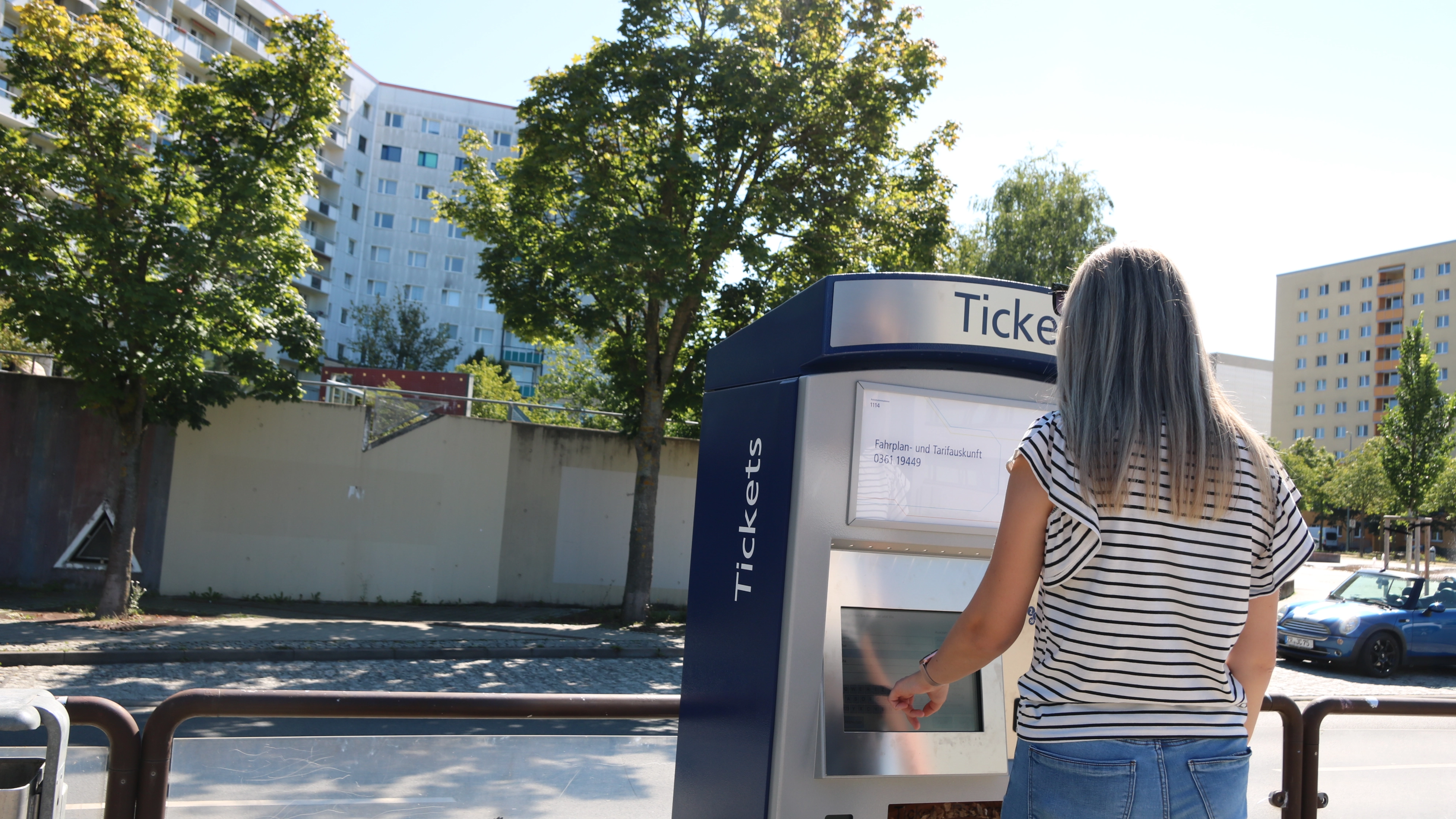 Eine Frau bedient einen Fahrscheinautomaten an einer Haltestelle. Im Hintergrund sind Bäume zu sehen.