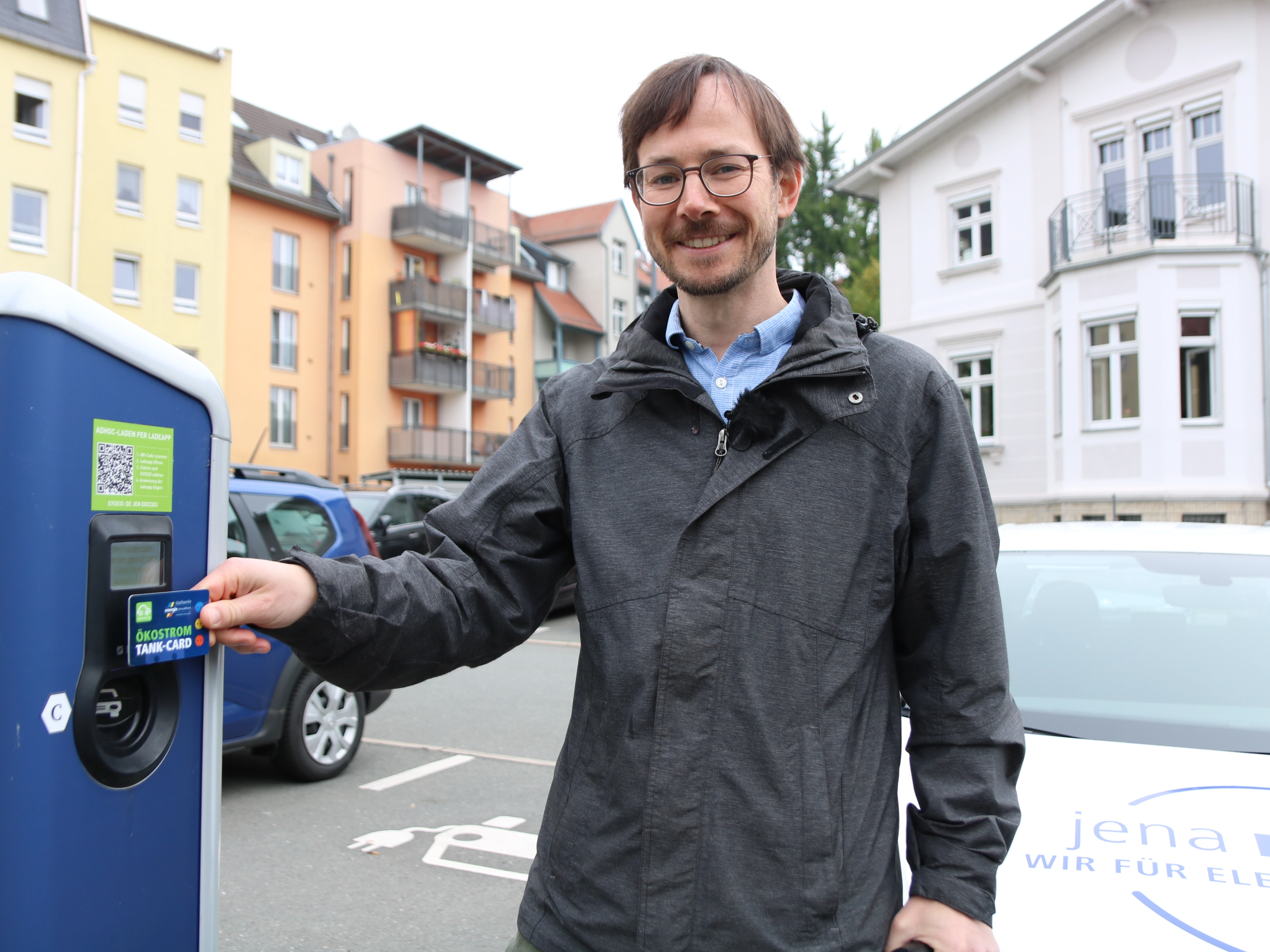 Ein Mann steht neben einer E-Ladesäule, sein Auto ist im Hintergrund sichtbar.