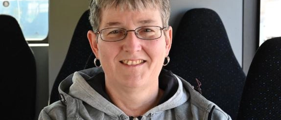Eine Frau mit Brille sitzt in einem Bus und lächelt in die Kamera. 