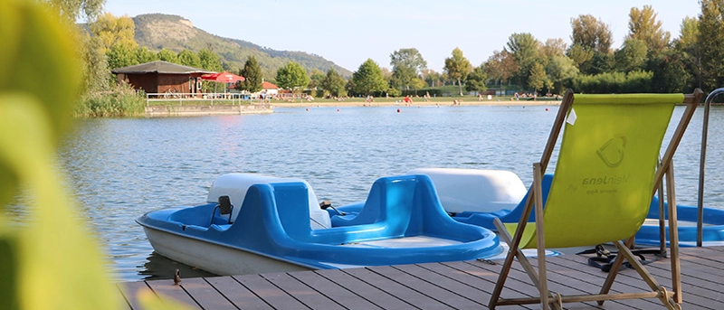 Eine Liegestuhl steht auf einem Steg, Tretboote befinden sich im Wasser 