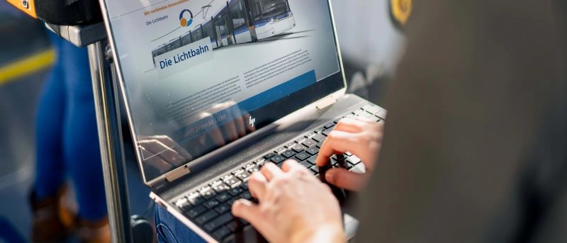 Person sitzt im Bus und hat einen Laptop auf dem Schoß. Auf diesem ist eine Website zur Lichtbahn zu sehen.