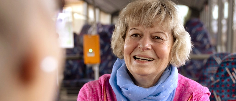 Eine blonde Frau sitzt im Bus und lächelt an der Kamera vorbei eine Person an, die im Anschnitt von hinten zu sehen ist.