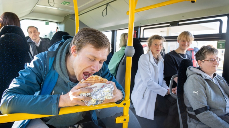 Ein Mann sitzt im Bus und isst einen Döner. Rundherum sitzen Fahrgäste und schauen zu dem Mann.